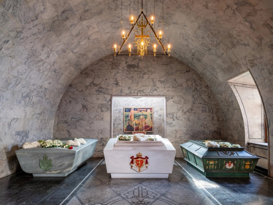 Three sarcophagi beneath a vaulted marble ceiling in the mausoleum, with a chandelier above and an altarpiece in the background