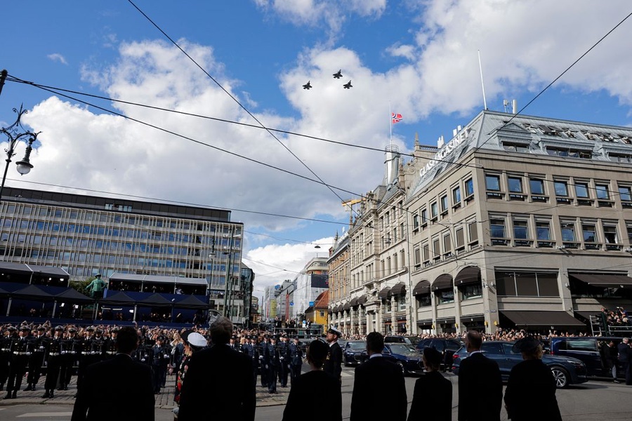 Four F-35 fighter jets fly over central Oslo in formation, with a Norwegian flag at half mast on a building below