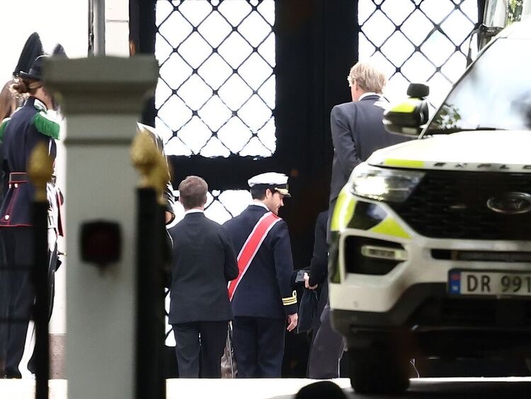 A man in uniform with a red sash walks in through the Palace gate together with a man in a black suit, with guards in the background