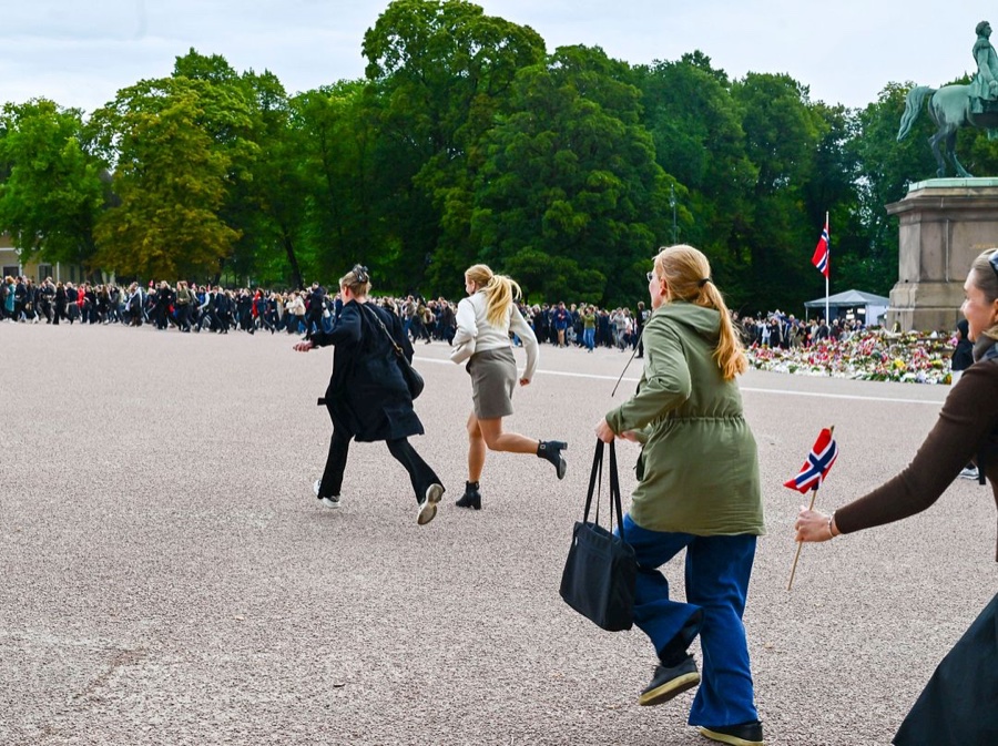 Three people run across the gravel of the Palace Square towards the Palace, one holding a Norwegian flag, with the sea of flowers and the crowd behind