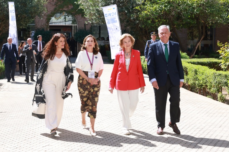 Queen Sofía walks along a sunlit path with three others, congress banners in the background