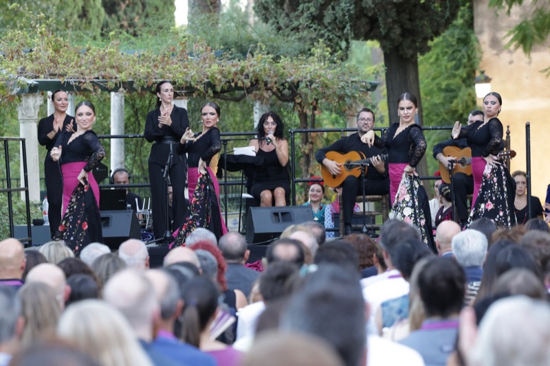 Flamenco dancers in black dresses with pink skirts perform on an outdoor stage in front of a full audience