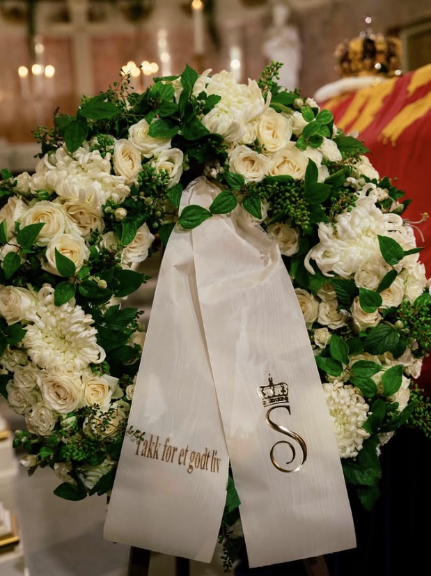 Close-up of a white wreath of roses and chrysanthemums, the ribbon bearing the words Takk for et godt liv and a crowned S monogram