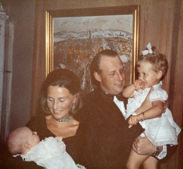 Older private colour photograph in which Harald and Sonja stand in front of a painting, he holding a small child and she holding a baby in a white gown