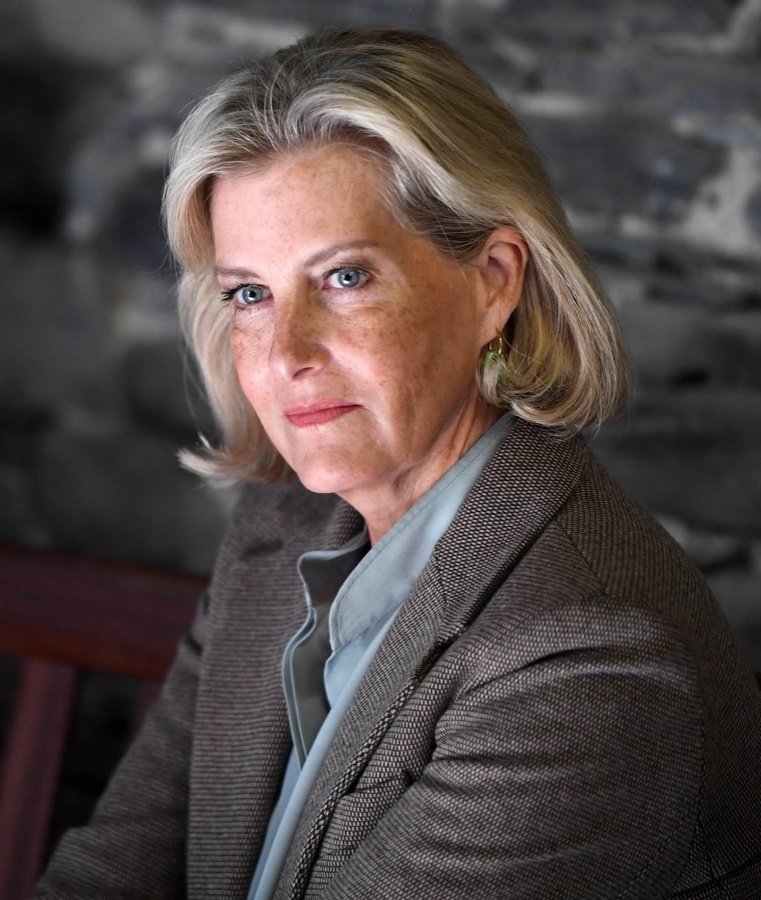 Portrait of the Duchess of Edinburgh in a brown blazer and pale blue blouse in front of a stone wall