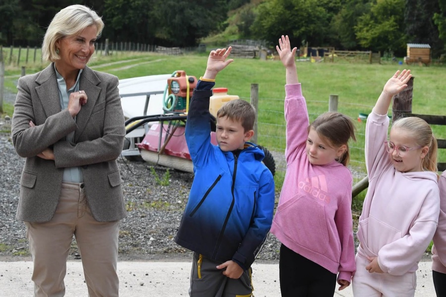 The Duchess of Edinburgh stands with her arms folded, smiling, as three schoolchildren raise their hands in a farmyard