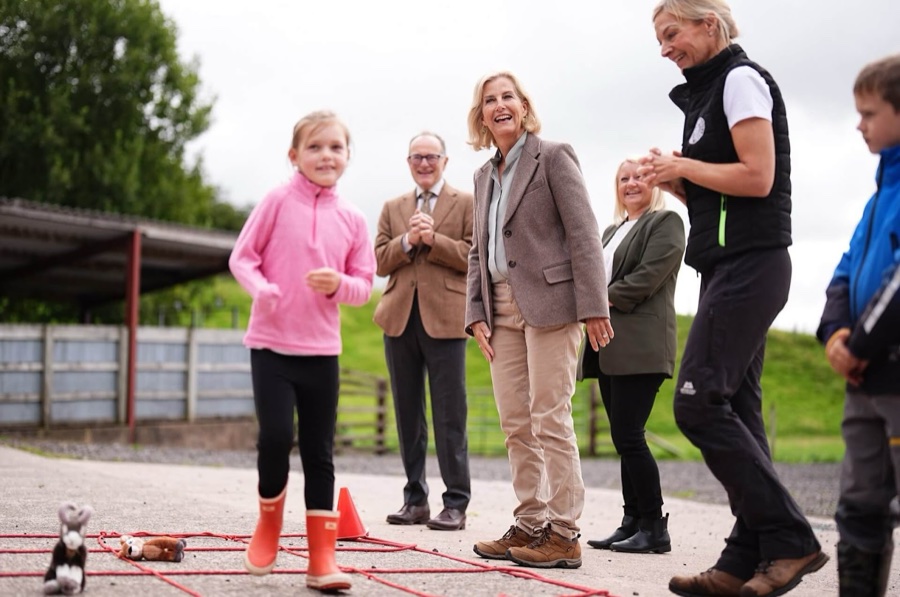 The Duchess of Edinburgh laughs as a girl in a pink fleece runs through a course of ropes and cones in the farmyard