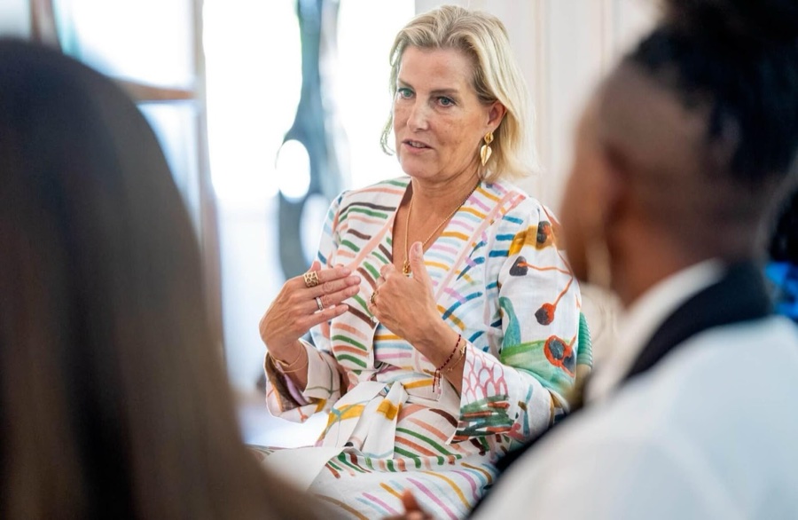 The Duchess of Edinburgh sits talking with her hands in motion, seen between the shoulders of two listeners