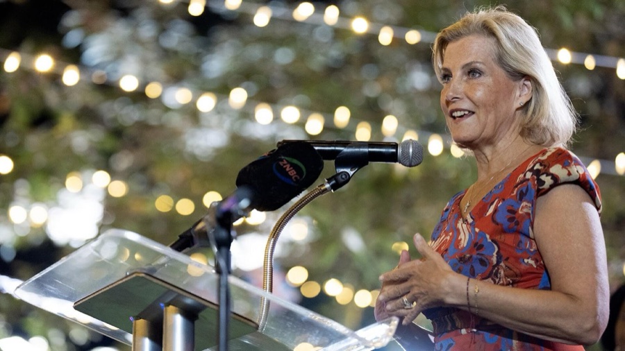 The Duchess of Edinburgh speaks from behind a glass lectern with microphones, with string lights in the background