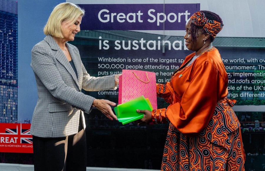 The Duchess of Edinburgh receives a pink gift bag and green folders from a woman in orange, in front of a screen with British campaign material