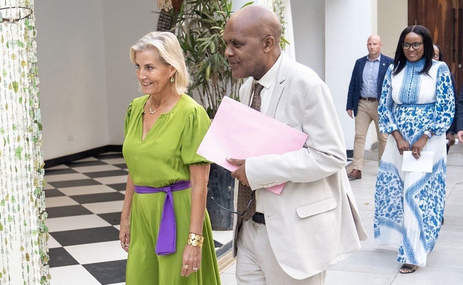 The Duchess of Edinburgh in a green dress with a purple belt walks with a man in a light suit carrying a pink folder
