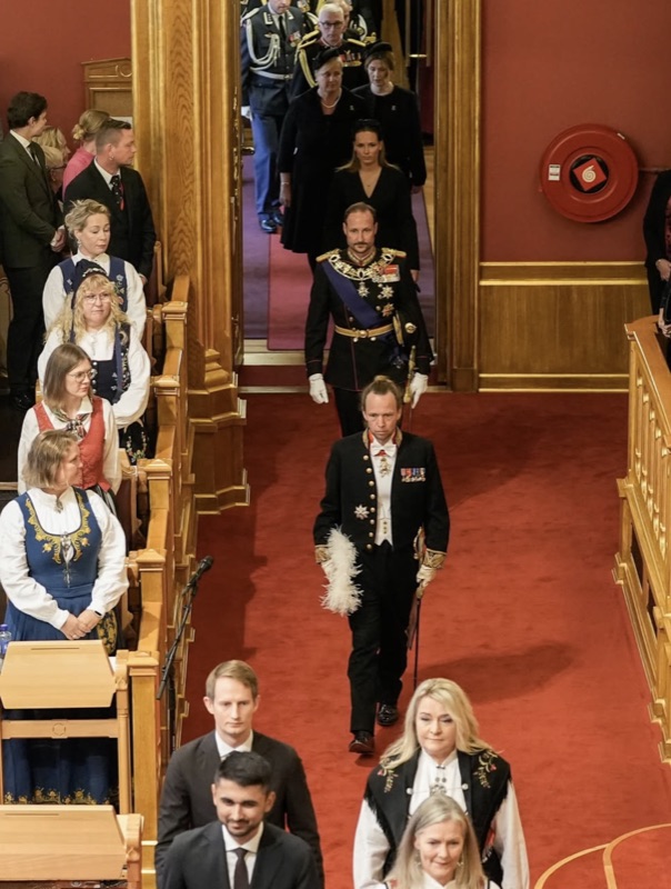 The procession entering the chamber, the King behind an officer, with members in national costume standing along the benches