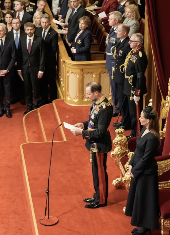 King Haakon reads the speech from a sheet of paper at the microphone, with Crown Princess Ingrid Alexandra standing by the throne