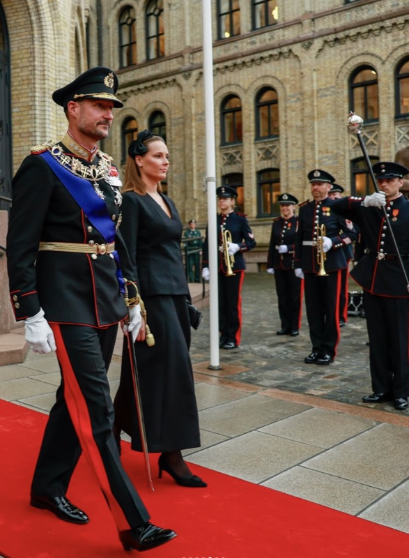 King Haakon and Crown Princess Ingrid Alexandra walk along a red carpet towards the parliament building, with musicians of the Royal Guard behind them