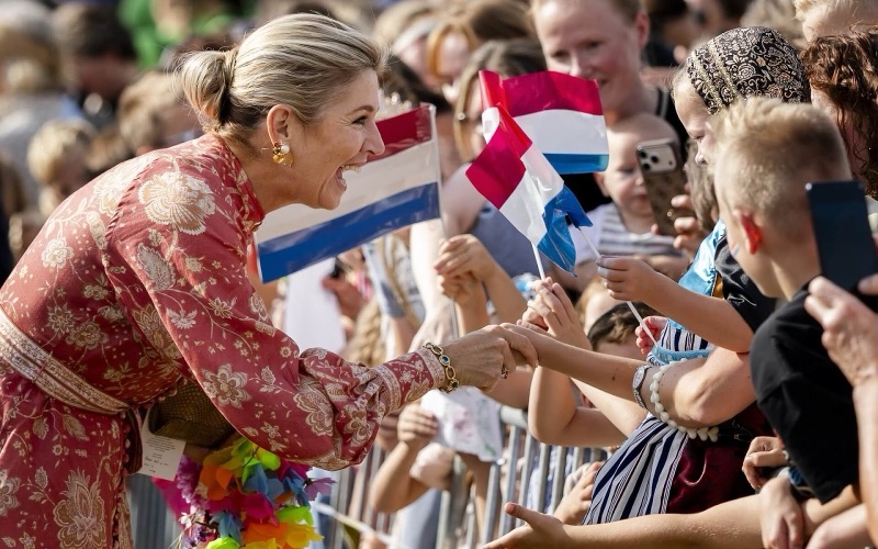 Dronning Máxima håndhilser på et barn over sperregjerdet, omgitt av nederlandske flagg