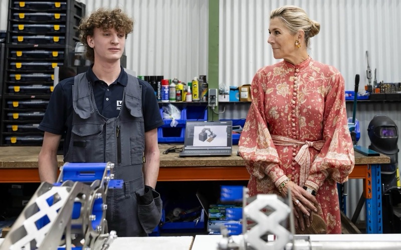 Dronning Máxima står i et verksted sammen med en ung mann i arbeidstøy, med robotdeler i forgrunnen
