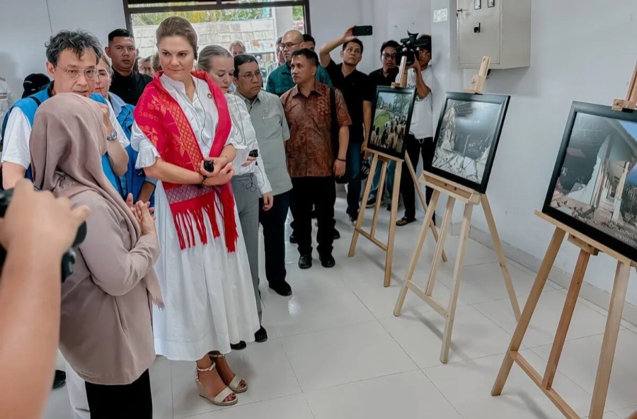 Crown Princess Victoria in a red scarf looks at framed photographs of damaged buildings displayed on easels