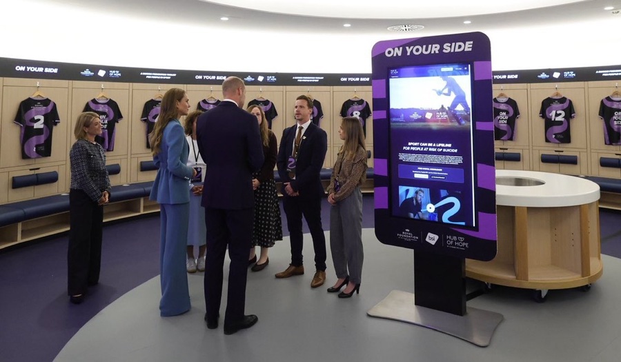 The Prince and Princess stand in the dressing room in front of a screen reading On Your Side, surrounded by shirts hanging on pegs