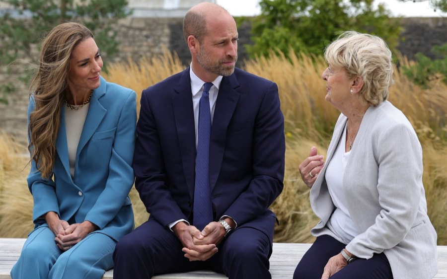 Prince William and Princess Catherine sit on a bench outdoors in conversation with a woman